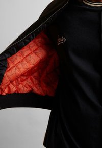 Blouson aviateur noir avec fermeture éclair à l'avant, doublure matelassée orange avec motifs géométriques, et col côtelé. Logo "Starter" brodé sur la poitrine.