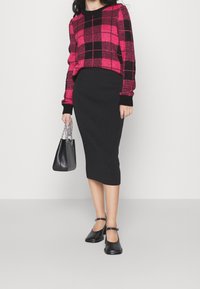 Rosa-schwarz karierter Pullover mit schwarzem, geripptem Midirock. Schwarze Schuhe mit Knöchelriemen und eine kleine schwarze Handtasche mit Kettenhenkel.