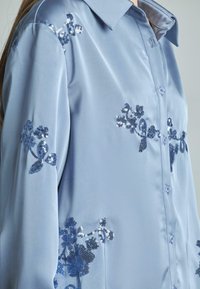 Camicia a maniche lunghe in satin azzurro chiaro con imbottiture di paillettes floreali. Presenta un colletto e chiusura con bottoni. Tessuto morbido.