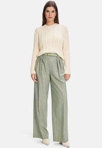 Cream cable-knit sweater and light green pleated trousers with a matching belt. Model stands, wearing neutral heeled shoes.