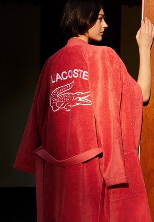 Red terrycloth bathrobe featuring a large white embroidered "LACOSTE" logo and crocodile design on the back; belt at the waist.