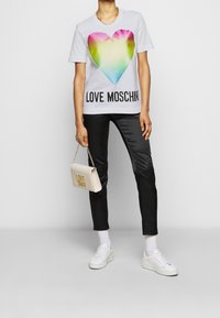 Bílý tričko s velkým barevným srdcem a textem "LOVE MOSCHINO". Vytvořeno s černými přiléhavými kalhotami, bílými teniskami a malou kabelkou.