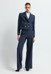 Vrouw in een marineblauwe pinstripe cropped dubbel-breasted blazer met gouden knopen, bijpassende wijde broek en zwarte hakken.