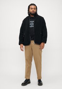 Sort fleecejakke med høj krave, lynlåslukning foran og to sidelommer, båret over en grafisk t-shirt, parret med lysebeige bukser og sorte sko.