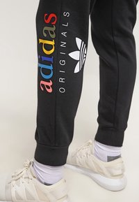 Nohy v černých teplákách Adidas Originals s barevným logem a bílými teniskami stojí na světlé podlaze.