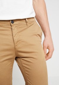 Chino in cotone beige con una superficie liscia, dotati di tasche frontali, chiusura con bottone e dettagli di cucitura vicino alla vita.
