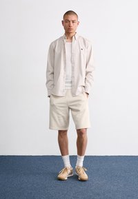 Beige long-sleeve shirt over a white tank top, paired with beige shorts. Light brown sneakers and white socks complete the outfit.