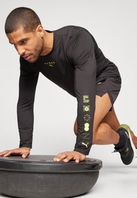 Schwarz langärmliges Sportshirt mit gelben Grafiken, kombiniert mit Shorts, schwarzem Schuhwerk und im Gleichgewicht auf einem grauen Trainingsgerät.