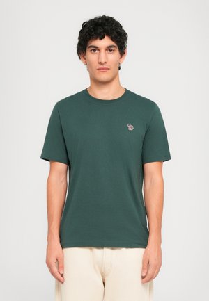 Ung mand med krøllet hår iført en mørkegrøn t-shirt med et lille farverigt zebra-logo på venstre bryst og beige bukser.