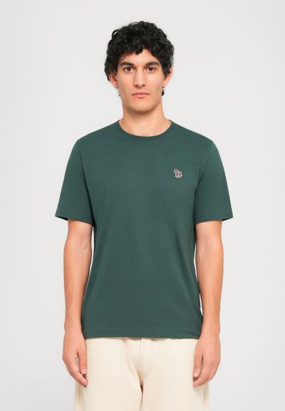 Jeune homme aux cheveux bouclés portant un t-shirt vert foncé avec un petit logo de zèbre coloré sur la poitrine gauche et un pantalon beige.