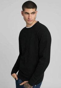 Pull en maille noire avec col rond, manches longues et poignets côtelés. Porté par-dessus une chemise blanche, associé à un jean bleu. Design simple et uni.