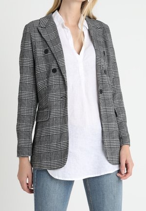 Femme portant un blazer à carreaux noir et blanc par-dessus une chemise blanche et un jean bleu, debout devant un fond clair uni.