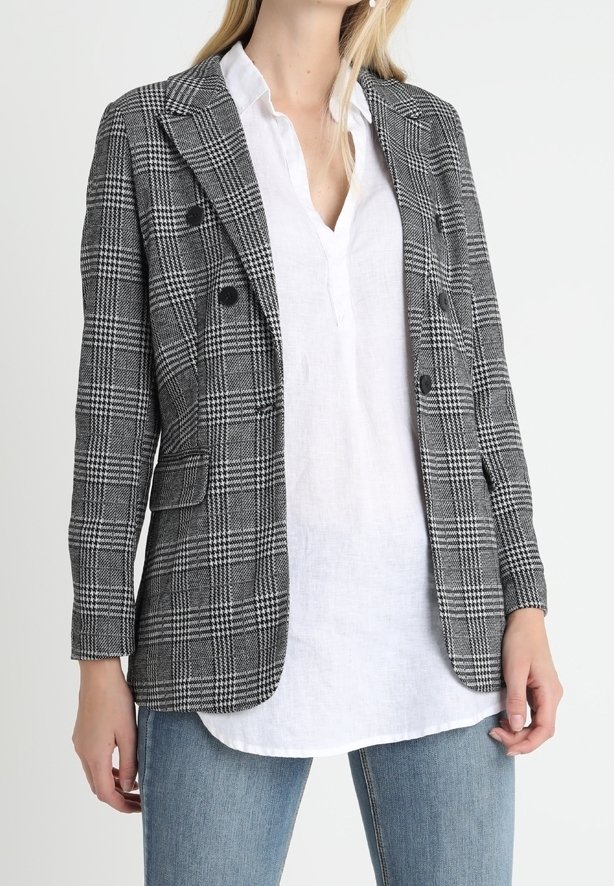 Femme portant un blazer à carreaux noir et blanc par-dessus une chemise blanche et un jean bleu, debout devant un fond clair uni.