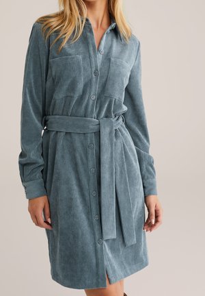 Kvinde iført en slået, bæltebeklædt, langærmet blå fløjlskjortekjole med brystlommer mod en ensfarvet baggrund.