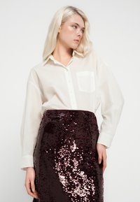 Chemise blanche surdimensionnée avec une poche avant, associée à une jupe crayon bordeaux à sequins, présentant une surface brillante et texturée ainsi qu'une silhouette ajustée.