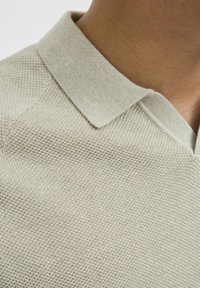 Lys beige polo skjorte med en teksturert strikkemønster, med en skarp krage og en V-formet halslinje, som viser fine detaljer.