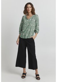 Blusa floreale verde con maniche a sbuffo, scollo a v; abbinata a pantaloni neri larghi. Accento di sandali neri con cinturini intrecciati.