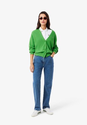 Groene wollen cardigan met V-hals en knoopsluiting; gecombineerd met een wit overhemd met kraag, blauwe spijkerbroek en witte sneakers. Accessoires zijn een zonnebril.