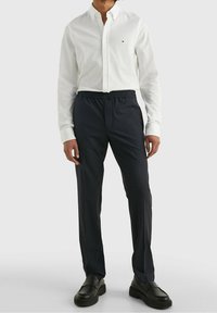 Camicia bianca con bottoni in cotone, abbinata a pantaloni elasticizzati navy scuro con motivo. Il modello indossa scarpe nere slip-on.