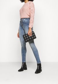 Suéter de punto de color rosa claro con patrón de hojas, jeans ajustados de cintura alta azules y un bolso de mano acolchado negro. Botines negros con tacón.