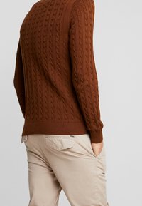 Pull en tricot torsadé marron avec poignets et ourlet côtelés, associé à un pantalon beige clair présentant une poche arrière et des détails subtils.