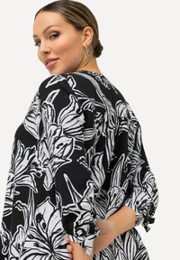 Sort bluse med hvidt blomsterprint, lange ærmer med bånd ved manchetterne, afslappet pasform, let stof, bagview der viser læg og halsudskæring.