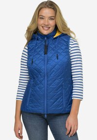 Modrá prošívaná vesta s vysokým límcem a žlutým vnitřkem kapuce. Má plný zip a dvě přední kapsy, nošena přes pruhované tričko.