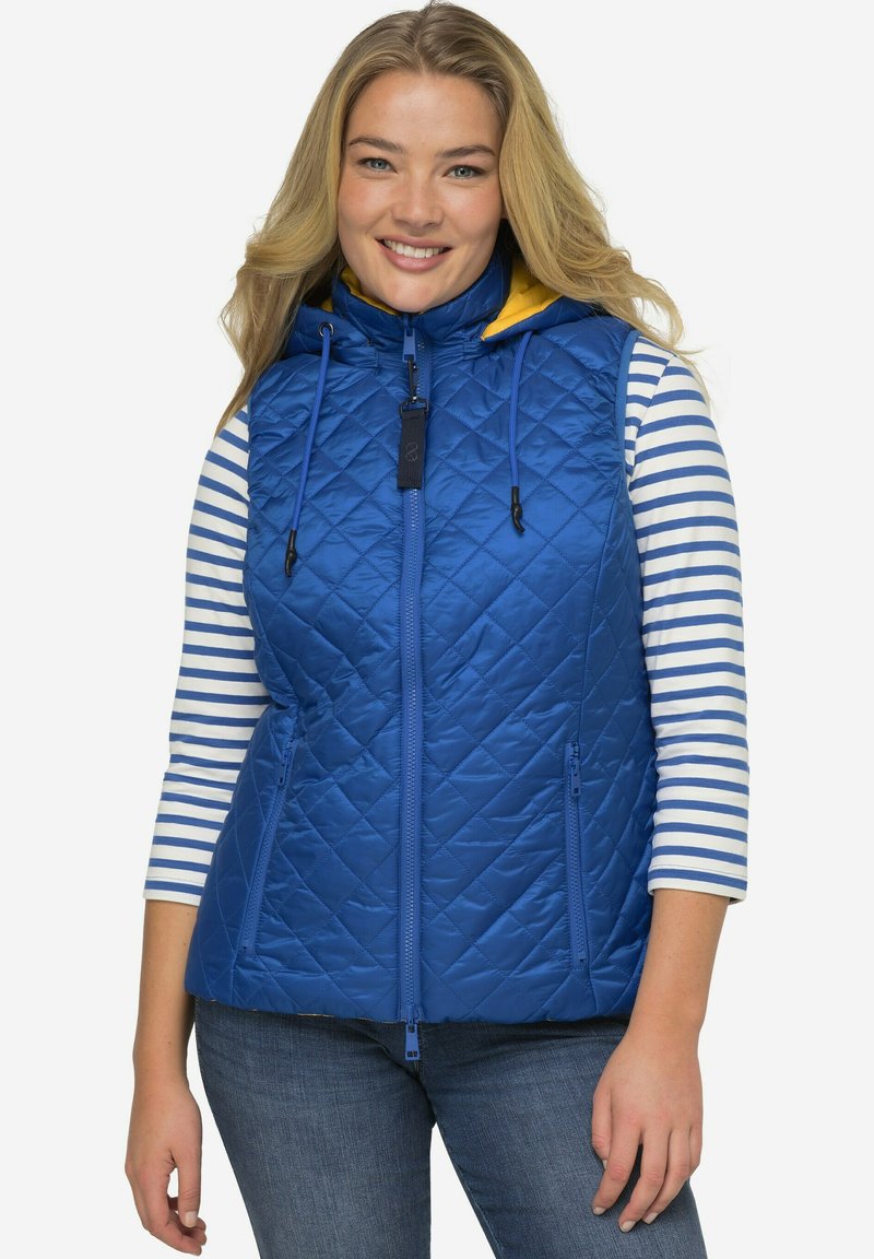 Modrá prošívaná vesta s vysokým límcem a žlutým vnitřkem kapuce. Má plný zip a dvě přední kapsy, nošena přes pruhované tričko.