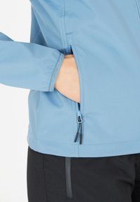 Veste zippée bleu clair avec une poche latérale. Le tissu semble lisse, avec un traitement déperlant. Le zip a des tirettes et des coutures noires.