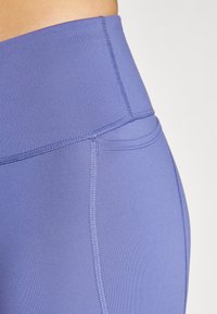 Leggings taille haute en tissu violet clair, dotés de coutures plates et d'une texture lisse avec une poche dissimulée dans la ceinture.