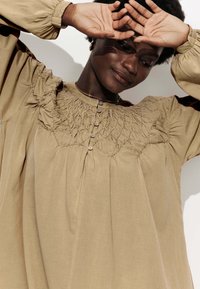 Blouse beige à manches longues avec smocks texturés et détails de boutons au niveau du col. Tissu lisse avec des manches froncées, coupe décontractée.
