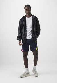 Fekete nylon zipzáras dzseki, fehér póló, tengerészkék sort vibráló rózsaszín és zöld részletekkel, valamint krémszínű sneakerek texturált talppal.