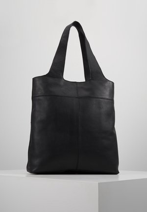 Czarna skórzana torba typu tote z szerokimi uchwytami, prostym wzorem i widocznym przeszyciem, stojąca pionowo na białym postumencie na jednolitym tle.