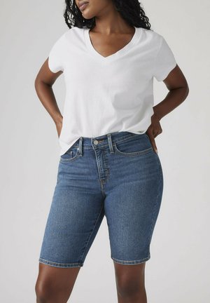 Vrouw draagt een wit T-shirt met V-hals en blauwe denim shorts tot op de knie, handen in haar zij, neutrale achtergrond.