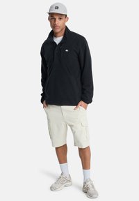 Sudadera de forro polar negra con media cremallera y un bolsillo en el pecho, combinada con pantalones cortos de cargo beige y zapatillas grises, sobre un fondo blanco.