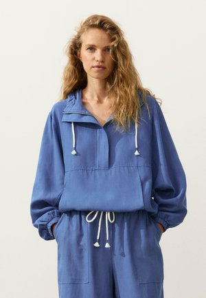 Kobieta o falowanych blond włosach, ubrana w luźny niebieski sweter i dopasowane spodnie z białymi sznurkami, stojąca z rękami w kieszeniach.