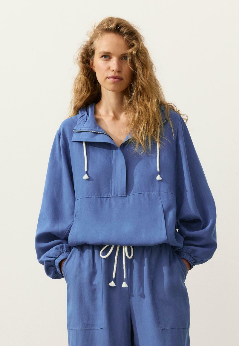 Femme aux cheveux blonds ondulés portant un pull bleu ample et un pantalon assorti avec des cordons blancs, debout les mains dans les poches.