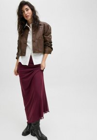 Chaqueta de cuero marrón corta con dos bolsillos delanteros, camisa blanca de botones y falda larga burdeos. Botines negros completan el conjunto.