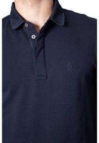 Jūros mėlynos spalvos polo marškiniai, pagaminti iš tekstūrinio medvilnės, su sagų užtrauktuku ir subtiliu logotipu kairėje krūtinės pusėje. Yra klasikinis apykaklė.