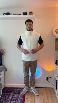 Vesta albă fără mâneci peste o cămașă neagră, asociată cu pantaloni cargo bej și adidași gri. Țesătură fină, închidere cu fermoar și buzunare vizibile.
