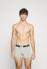 Bóxer gris con cinturilla elástica negra que presenta una marca blanca. Estilo ajustado con una textura suave. El modelo muestra la vista frontal.