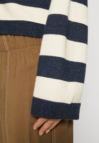 Marinblå och krämrandig tröja med en texturerad stickning, med breda manschetter. Kombineras med tan-färgade byxor som har detaljerad resårmidja.