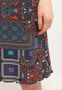 Main reposant sur une jupe avec des motifs cachemire floraux et géométriques multicolores complexes en bleu, orange, rouge, noir et blanc.
