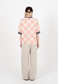 Persoană îmbrăcată într-o cămașă cu mânecă scurtă, în carouri roz și alb, pantaloni largi bej și o șapcă neagră de baseball, văzută din spate pe un fundal alb.