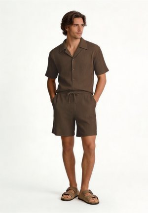 Homme debout les mains dans les poches, portant une chemise marron à manches courtes boutonnée, un short assorti et des sandales beiges sur fond blanc.