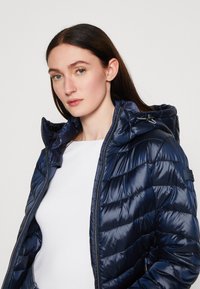 Doudoune marine avec une texture brillante et matelassée et une capuche détachable. Équipée d'une fermeture éclair et d'une patch de marque sur la manche.