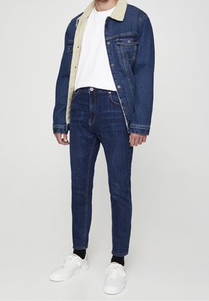 Chaqueta de denim con cuello de borreguillo beige, camiseta blanca, jeans de corte ajustado azul oscuro y zapatillas blancas. Atuendo casual con texturas contrastantes.