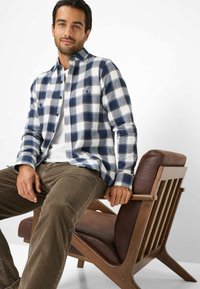 Camicia di flanella a quadri blu e bianchi da uomo con due tasche sul petto, abbinata a pantaloni in corduroy marroni testurizzati. Persona seduta su una sedia di legno.