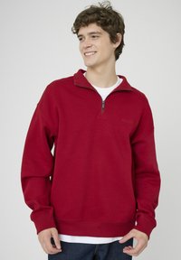 Sweat-shirt rouge avec col à demi-fermeture éclair, poignets et ourlet côtelés, présentant une texture de tissu lisse et un petit logo sur la poitrine.