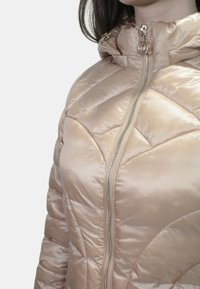 Giacca beige imbottita con motivo a trapunta, colletto alto e zip frontale. Presenta un tessuto lucido e un cursore della zip metallico.